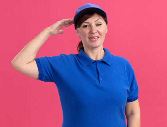 微笑身穿蓝色制服 头戴鸭舌帽的中年女送货员站在粉红色的墙上 面带微笑 自信地致意年龄敬礼站立