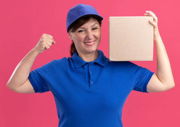 兴奋身穿蓝色制服 头戴鸭舌帽的中年女送货员拿着纸板箱看着前面 高兴而兴奋地握紧拳头站在粉色的墙上年龄拳头递送
