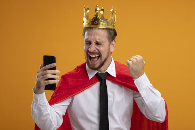 是的快乐的年轻超级英雄戴着领带 戴着王冠 看着手机 在橙色的背景上显示“是”的手势年轻快乐手势