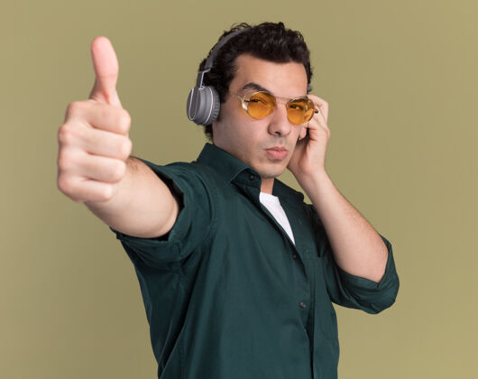 年轻人身穿绿色衬衫的年轻人戴着眼镜 戴着耳机 严肃地看着前面 竖起大拇指站在绿色的墙上衬衫脸拇指