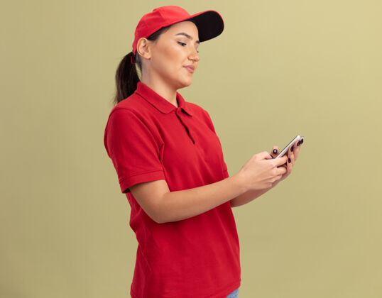 脸年轻的送货员穿着红色制服 戴着帽子 用智能手机看着它 脸上带着微笑站在绿色的墙上传递微笑年轻