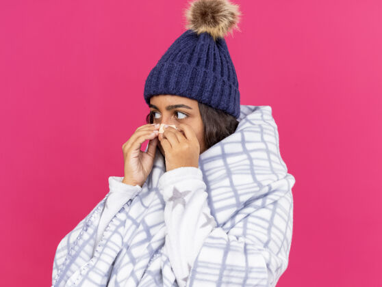 鼻子年轻生病的女孩一边看一边戴着冬天的帽子 围巾用格子布包着 鼻子上贴着膏药 隔离在粉红色的背景上年轻围巾石膏
