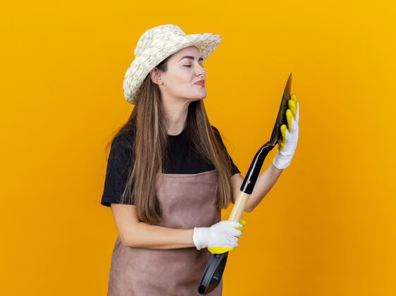制服高兴美丽的园丁女孩穿着制服 戴着园艺帽 戴着手套 拿着黑桃看着孤立的橙色背景黑桃园艺花园