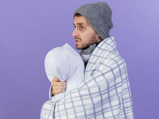枕头看着身边年轻的病夫戴着冬天的帽子 围巾裹着格子呢 抱枕孤立地放在紫色的背景上年轻拥抱帽子