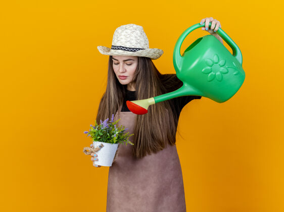 帽子美丽的园丁女孩穿着制服 戴着园艺帽 在花盆里浇花 用浇水罐隔离在橙色的背景上园艺女孩浇水