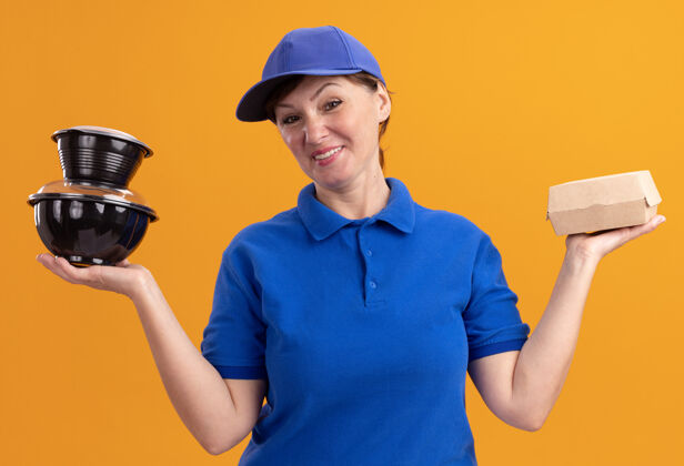 快乐身穿蓝色制服 头戴鸭舌帽的中年女送货员站在橘色的墙上 手里拿着食品包 开心而积极地看着前面女人抱着制服