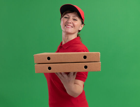年龄身穿红色制服 头戴鸭舌帽的中年送货妇女站在绿色的墙上 面带微笑地看着前面的比萨饼盒抱着送货站着