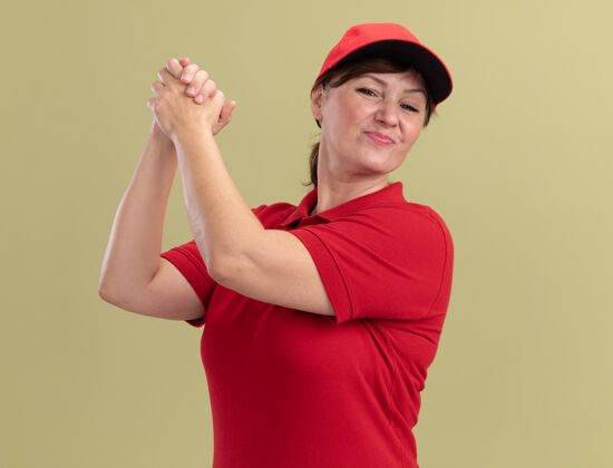 女人身着红色制服 头戴鸭舌帽的中年女送货员站在绿色的墙边 自信地手拉着手 做出团队合作的姿态手帽子立场