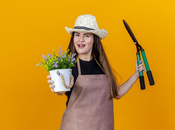 剪子困惑美丽的园丁女孩穿着制服 戴着园艺帽拿着剪子 拿着花盆里的花 对着橙色背景上孤立的镜头女孩帽子花园