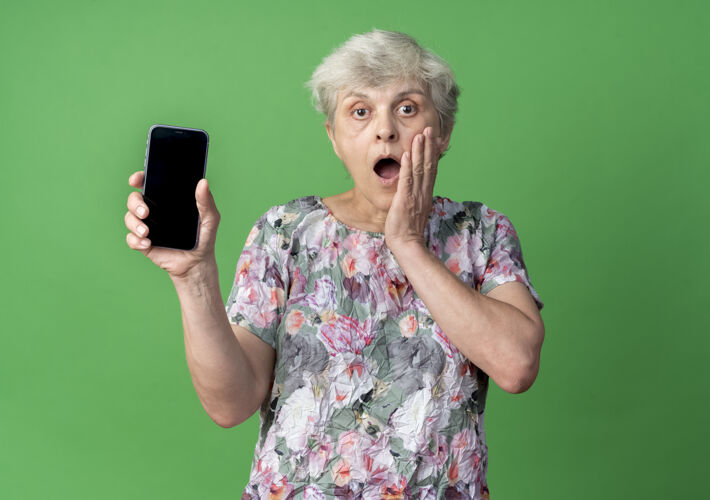 女人震惊的老妇人把手放在脸上拿着电话隔离在绿色的墙上老人绿色电话