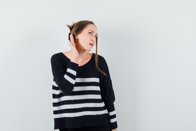 护理一个年轻的女人手举在耳边 试图听到一些穿着条纹针织衫和黑色裤子的声音 看起来很专注自然快乐焦点