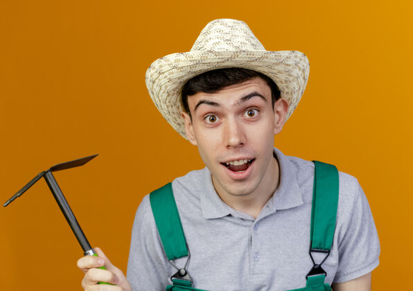 帽子惊讶的年轻男园丁戴着园艺帽拿着锄头耙看着园艺空间橙色