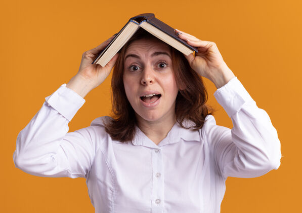 微笑身穿白衬衫的年轻女子头上举着一本打开的书 站在橘色的墙上困惑地微笑着抱着站着年轻