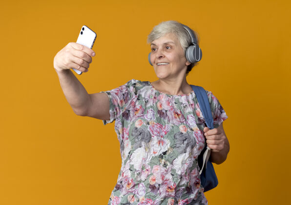 背包戴着耳机 背着背包 面带微笑的老妇人看着电话 独自在橙色的墙上自拍老人穿自拍