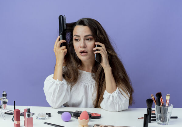 发夹震惊的美丽女孩坐在桌子旁 用化妆工具梳头 讲电话 看着紫色墙上孤立的一面电话桌子女人