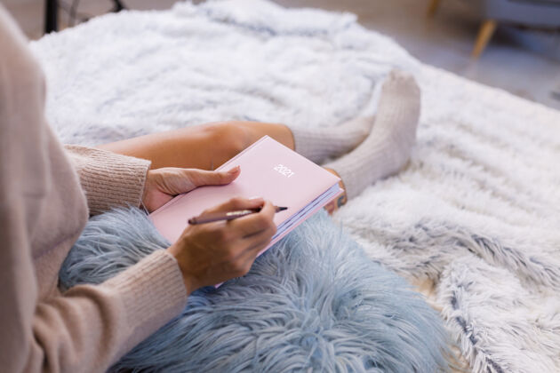 钢笔穿着羊毛袜子和毛衣的女人 粉色笔记本上写着2021 臀部有大纹身女人坐在卧室的家里的床上家庭作业毯子早晨