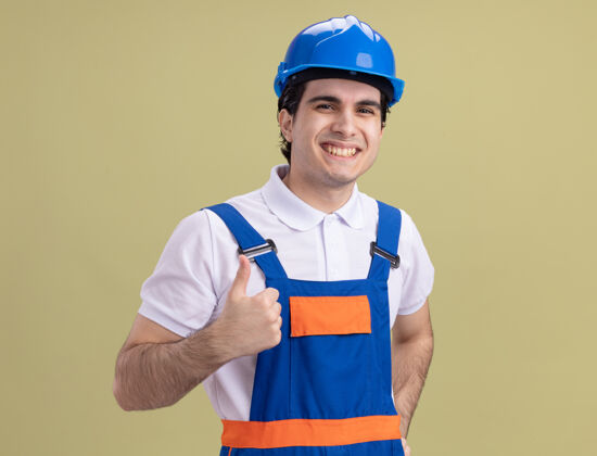建筑工人年轻的建筑工人身穿建筑制服 戴着安全帽 站在绿色的墙壁上 面带微笑 高高兴兴地竖起大拇指年轻头盔欢呼