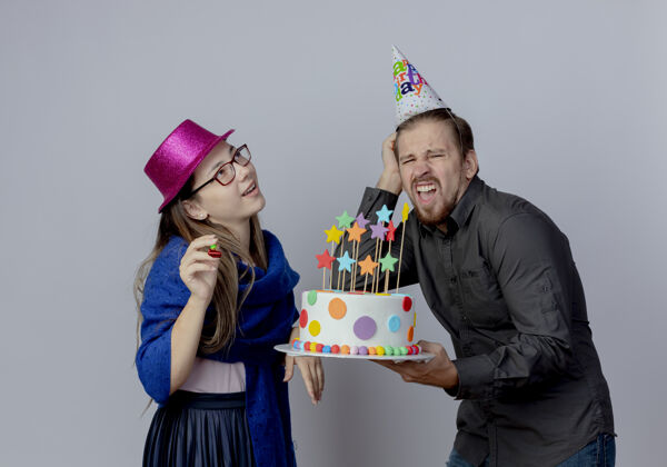 女人戴着粉红色帽子戴着眼镜的年轻女孩高兴地举着哨子抬头看 戴着生日帽的帅哥生气地拿着蛋糕 把手放在隔离在白墙上的头上粉色蛋糕穿