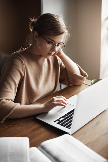 笔记本电脑戴着时髦眼镜的白人女企业家 目光集中 头靠在手上 盯着笔记本电脑屏幕 在笔记本和书本上做笔记 努力工作Wifi无线企业家