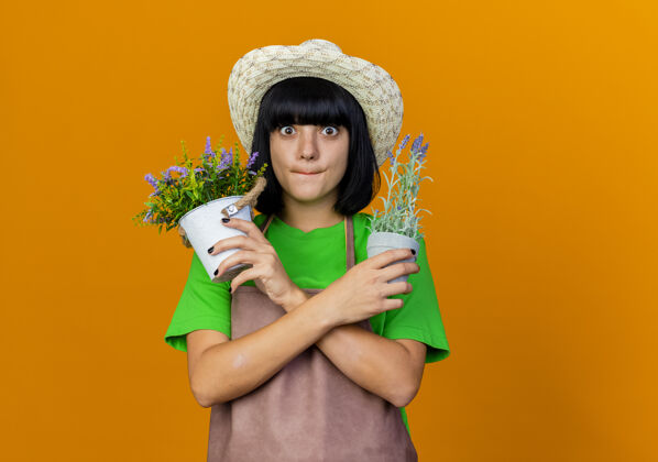 帽子焦虑的年轻女园丁穿着制服 戴着园艺帽 双手交叉握着花盆年轻橙色园艺