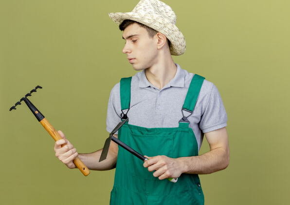 绿色自信的年轻男园丁戴着园艺帽 拿着耙子和锄头耙子看着旁边年轻空间穿