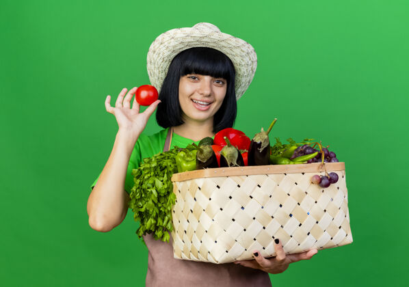 举行快乐的年轻女园丁穿着制服 戴着园艺帽 手里拿着菜篮子和西红柿空间帽子花园