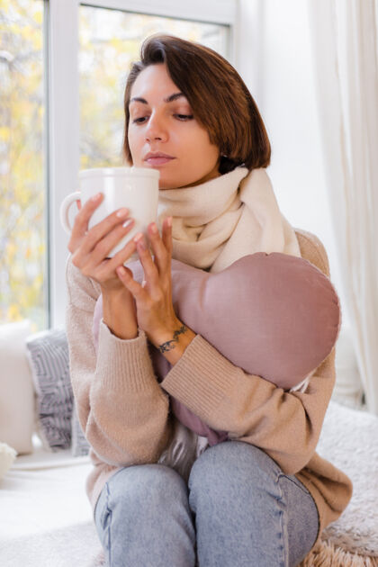 杯子坐在窗台上喝着热茶喝着咖啡 穿着毛衣和白围巾的女人的温馨肖像季节积极放松