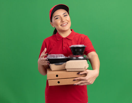 披萨身穿红色制服 头戴鸭舌帽的年轻女送货员手持披萨盒和食品包 面带微笑地站在绿色的墙上 看着前方站着送货微笑