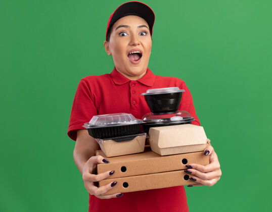 制服年轻的送货员身穿红色制服 头戴鸭舌帽 手里拿着披萨盒和食品包 站在绿色的墙边惊讶地看着前面拿着帽子送货