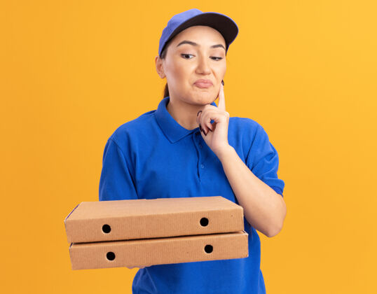 拿着站在橘色的墙上 一位身穿蓝色制服 戴着帽子 拿着披萨盒 脸上带着沉思表情的年轻女送货员看着披萨盒送货制服帽子