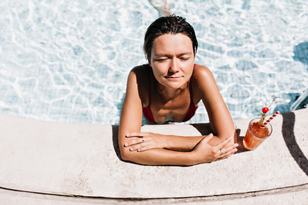 欧洲黑发女孩在游泳池里晒太阳晒黑的女人在水里用鸡尾酒冷却成人泳装阳光