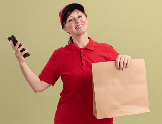女人快乐的中年送货妇女 穿着红色制服 戴着帽子 手里拿着纸包和智能手机 面带微笑 自信地站在绿色的墙边快乐递送自信