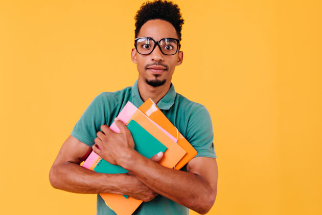 朋友穿着鲜艳t恤的非洲学生摆出惊讶的表情戴眼镜的黑人男孩拿着书站着看学术笔记本大学