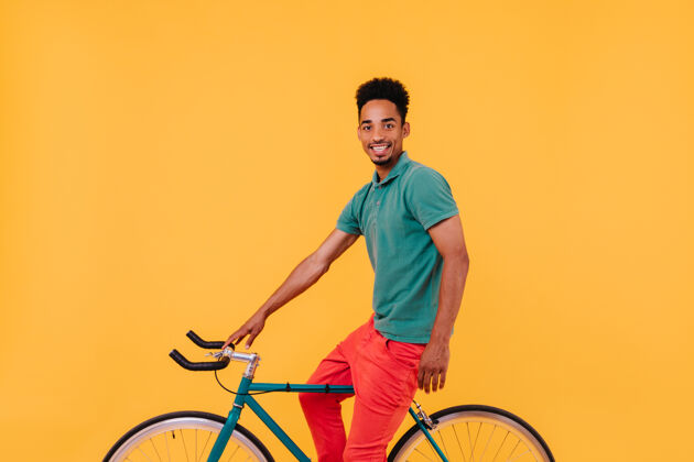 休闲优雅的男自行车手微笑着摆姿势有兴趣的黑发男子坐在黄墙附近的自行车上微笑时髦兴奋