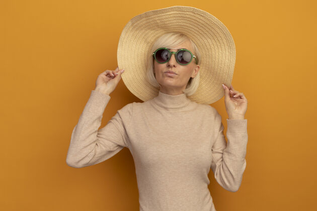 女人自信的金发斯拉夫女人 戴着沙滩帽 戴着太阳眼镜 站在橙色的沙滩上时尚自信太阳