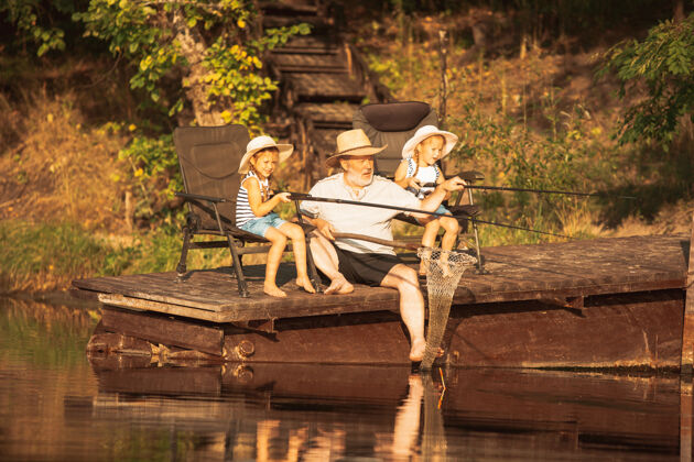 爱好可爱的小女孩和他们的爷爷在湖边或河边钓鱼在夏天的日落时分 在靠近水和森林的码头上休息家庭 娱乐 童年 自然的概念世代孙女女孩