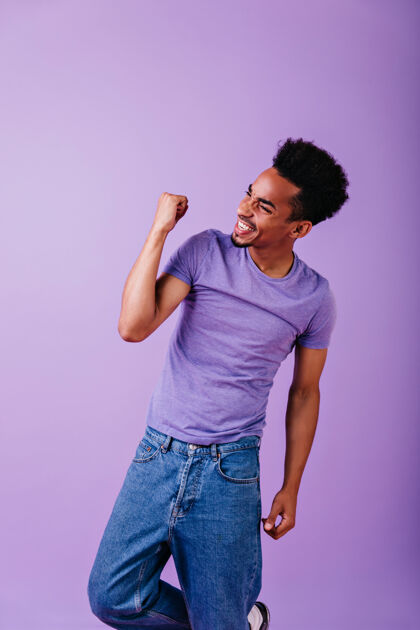 美国快乐帅气的男模穿着蓝色牛仔裤表达积极的情绪室内照片中的非洲短发小伙子穿着紫色t恤人迪斯科人