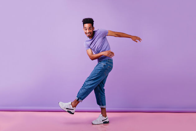 移动快乐的非洲男人带着真诚的微笑跳舞的全幅照片穿着牛仔裤和紫色t恤的男模玩得很开心家伙肖像自信