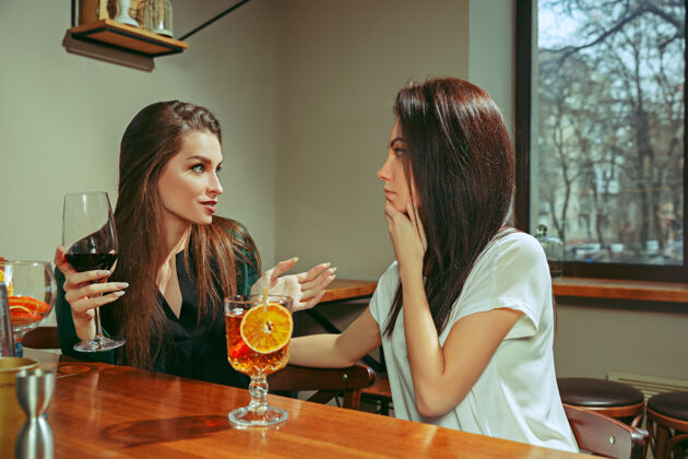 手女朋友在酒吧喝酒她们坐在一张摆着鸡尾酒的木桌旁她们穿着休闲服一起美丽女孩