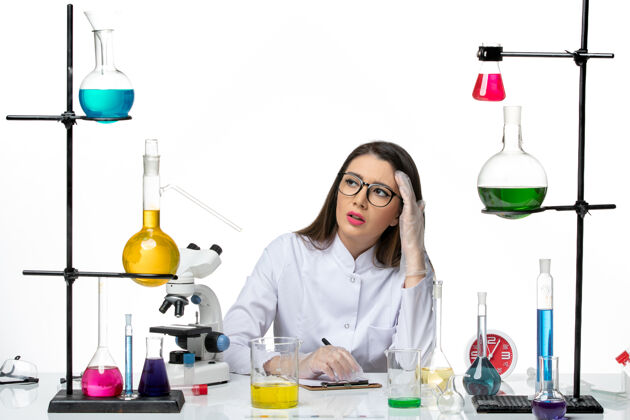 视图前视图穿着白色医疗服的女化学家坐在一个白色背景的科学大流行病毒冠状病毒实验室里 拿着溶液写笔记成人解决方案科学