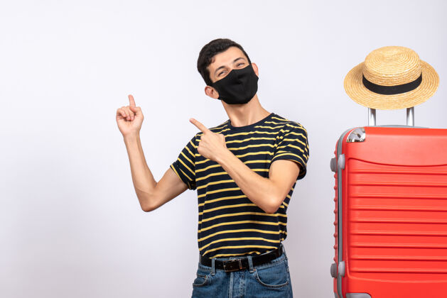 男人正面图：戴着黑色面具的快乐年轻游客站在红色手提箱旁边 指着后面站立成人肖像