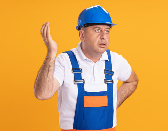 站立不知所措的白人成年建筑工人身着制服 站在那里举起手 看着橙色的一面人不知所措制服