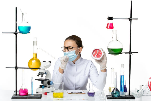 咳嗽前视图：身穿医疗服的女化学家 戴着面罩 手持红色时钟 在白色背景下咳嗽病毒实验室化学冠状病毒-飞溅研究病毒飞溅