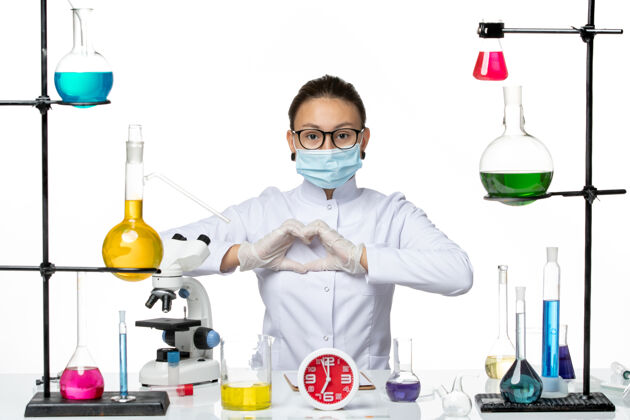 视图前视图：穿着医疗服的女化学家 戴着面罩 坐在白色办公桌上 拿着溶液 化学病毒实验室病毒-飞溅面罩实验室医疗