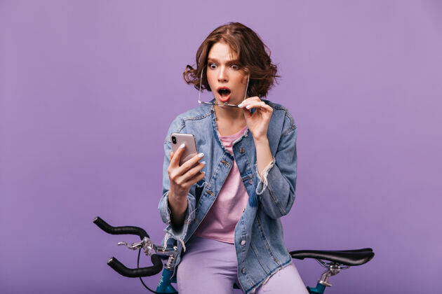 自行车穿着牛仔夹克的忧心忡忡的女孩看着手机屏幕室内镜头是一位迷人的白人女士坐在自行车上 拿着智能手机 表达着惊奇车辆运动员休闲