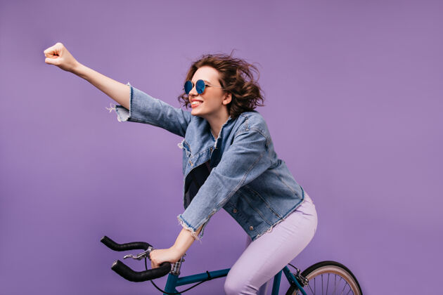 女士穿着牛仔夹克的自信女孩骑在自行车上挥舞着手一张充满灵感的戴眼镜的年轻女士坐在自行车上的室内照片女士骑车人微笑