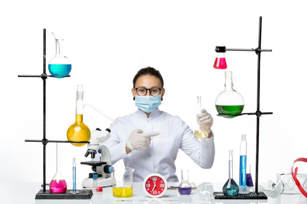 桌子前视图：穿着白色医疗服的女化学家 在桌子前面戴着面罩 白色背景上有溶液化学家实验室病毒冠状病毒飞溅飞溅实验化学家