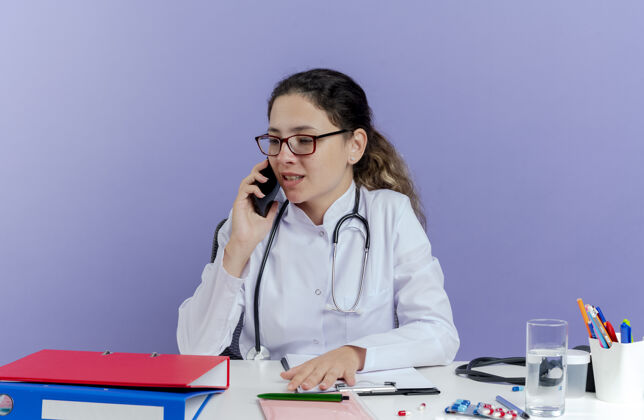 桌子高兴的年轻女医生穿着医用长袍和听诊器坐在办公桌旁 拿着医疗工具一边看一边讲电话医疗医生长袍