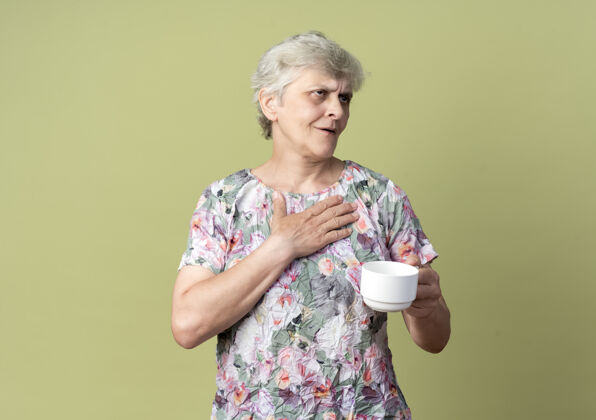 杯子高兴的老妇人把手放在胸前 举着杯子看着橄榄绿墙上孤立的一面老年人手橄榄色