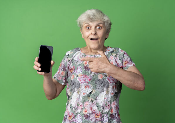 老人兴奋的老妇人拿着电话指着隔离在绿墙上的电话绿色点抱着